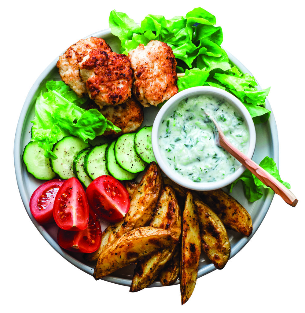 Fish skewers, baked potatoes, vegetables and yogurt greens sauce on dark background, top view